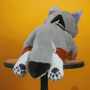 Peut inclure: Un jouet en peluche en forme de chat, vu de dos. Le chat est principalement gris avec des pattes blanches et des accents bruns. Les pattes ont des coussinets noirs. Le jouet est pos&eacute; sur un petit tabouret en bois sur fond jaune.