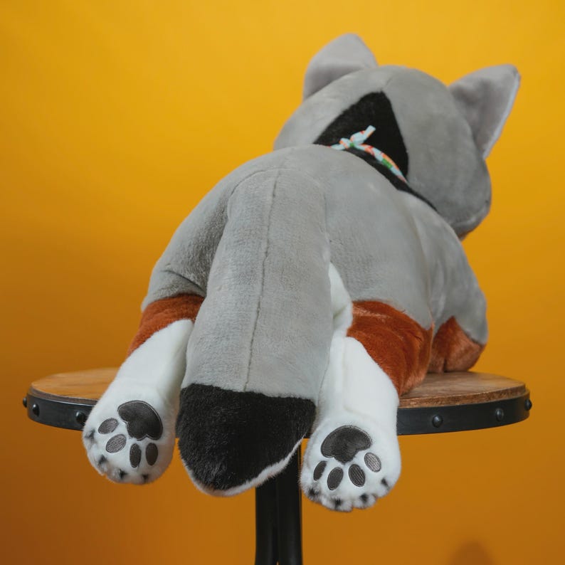Peut inclure: Un jouet en peluche en forme de chat, vu de dos. Le chat est principalement gris avec des pattes blanches et des accents bruns. Les pattes ont des coussinets noirs. Le jouet est pos&eacute; sur un petit tabouret en bois sur fond jaune.
