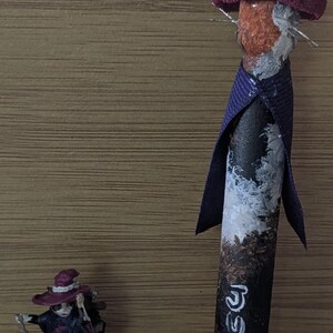 May include: A whimsical, handcrafted witch-themed decorative item. The larger piece is a painted wooden figure with a hat and ribbon. A smaller figurine of a witch stands nearby. The background is a wooden surface.