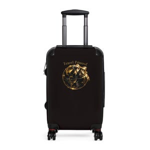 Può includere: Valigia nera con ruote e manico retrattile. La valigia presenta una grafica dorata di un globo con un fiocco e le parole "Travel Princess" in oro. La valigia ha ruote nere e un manico nero.