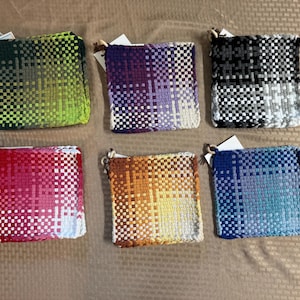 May include: Six square woven potholders featuring a grid-like pattern with color gradients. The set includes one green, one purple, one black and white, one red and pink, one orange and yellow, and one blue and teal potholder, each with a small loop.