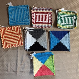 Puede incluir: Una colección de siete salvamanteles cuadrados tejidos en varios colores y patrones. Los salvamanteles presentan diseños en azul, rojo, blanco, verde, negro, gris y amarillo. Cada salvamanteles tiene un lazo para colgar.