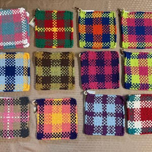 Op de afbeelding: Een collectie van elf vierkante geweven pannenlappen gerangschikt in drie rijen. Elke pannenlap heeft een verschillend veelkleurig ruitpatroon, gemaakt met dik garen in diverse combinaties van rood, blauw, groen, geel, roze en paars.