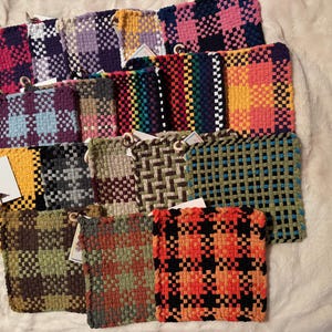 May include: A selection of woven potholders in a variety of colours and patterns. The potholders have a checkerboard design, with colours including pink, purple, green, orange, and blue. Each potholder has a loop for hanging.