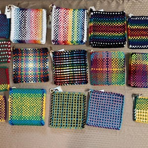 May include: A collection of fourteen square, woven potholders in various multicolored patterns, including stripes, plaids, and solid checkerboard designs. Each textile square features a small loop at one corner for hanging, showcasing diverse color combinations.