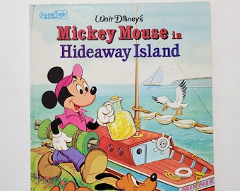 Op de afbeelding: Een vintage Golden Book getiteld Walt Disney's Mickey Mouse in Hideaway Island. De kaft toont Mickey Mouse in een roze shirt en pet, met een opgerolde mat en een glazen kan, terwijl Pluto een blauwe radio vasthoudt bij een rode boot.