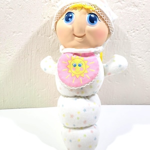May include: A vintage, plush doll with a smiling face, blue eyes, and blonde hair. It wears a white bonnet and a pink bib with a sun graphic. The doll's body is white with pastel star patterns, and it has small, soft arms.