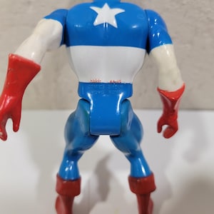 May include: A Captain America action figure, viewed from the back, showcasing a blue, white, and red color scheme. The figure has a white star on the chest, red gloves and boots, and the text "MARVEL COMICS GROUP HONG KONG" is visible.