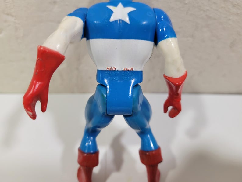 May include: A Captain America action figure, viewed from the back, showcasing a blue, white, and red color scheme. The figure has a white star on the chest, red gloves and boots, and the text "MARVEL COMICS GROUP HONG KONG" is visible.