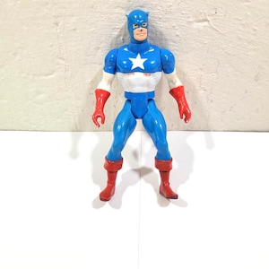 May include: A Captain America action figure in a classic superhero pose. The figure is predominantly blue, with a white star on the chest, red gloves and boots. The figure has a blue mask and a determined expression.