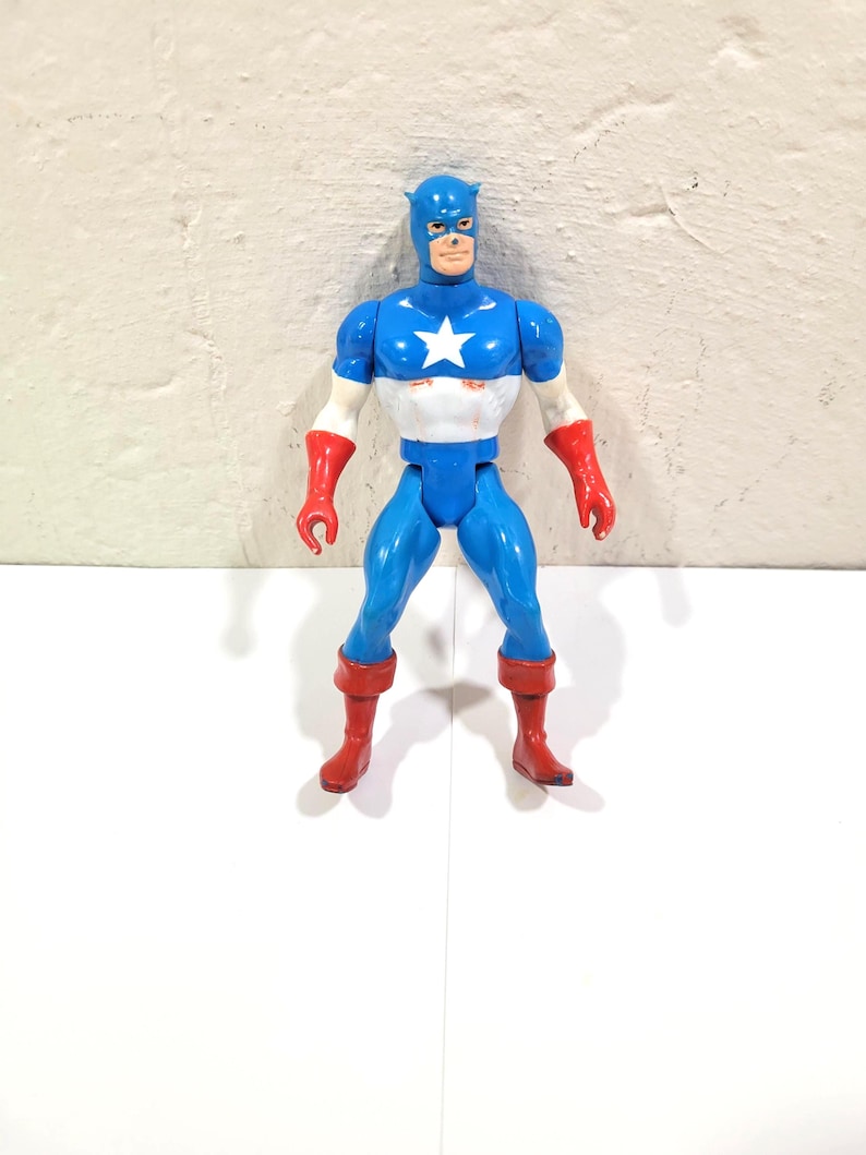 May include: A Captain America action figure in a classic superhero pose. The figure is predominantly blue, with a white star on the chest, red gloves and boots. The figure has a blue mask and a determined expression.