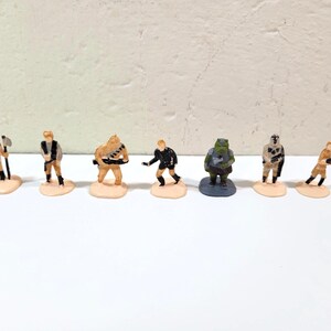 May include: A collection of seven small, painted figurines of various characters. Each figure stands on a small, light-colored base. The figures are in various poses and wear different outfits, with a variety of colors and designs.