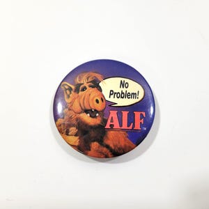 May include: A round button featuring the character ALF from the 1980s sitcom. The button has a blue background with ALF's image and the text "No Problem!" in a speech bubble. The word "ALF" is in red.