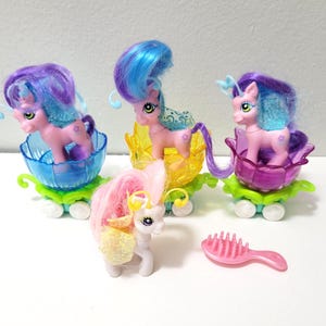 May include: Four toy ponies with colorful hair and flower-shaped carriages. The ponies are pink and white with various hair colors, including blue, purple, and pink. A pink comb is also present. The carriages are in shades of blue, yellow, and purple.