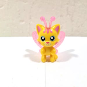 May include: A yellow cat figurine with pink butterfly wings and antennae. The cat has blue eyes and a pink stripe on its head. The figurine is standing on a white surface.