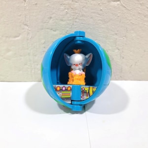 May include: A blue and green toy globe opens to reveal a small, silver-colored creature with large ears and a crown, seated on an orange platform. The globe has a detailed interior with colorful accents and a futuristic design.