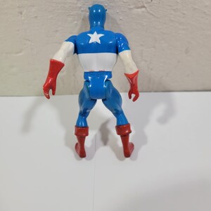 May include: A blue, white, and red Captain America action figure, viewed from the back. The figure has a blue helmet, a white star on the chest, and red gloves and boots. The figure is standing on a white surface.
