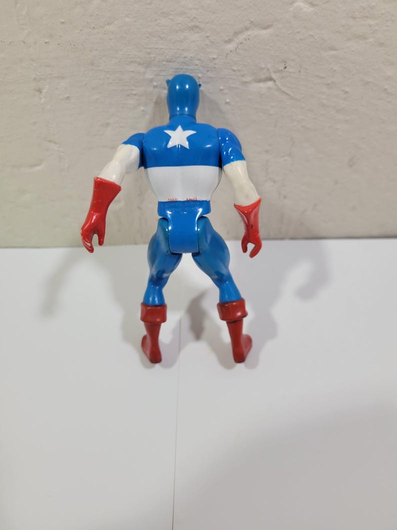 May include: A blue, white, and red Captain America action figure, viewed from the back. The figure has a blue helmet, a white star on the chest, and red gloves and boots. The figure is standing on a white surface.