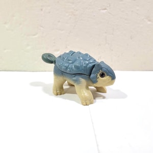 May include: A small plastic dinosaur figurine featuring a blue, armoured back with textured plates and a light beige underside. The creature has four short legs, a small tail, and a detailed head with dark eyes, resembling an ankylosaurus.