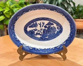 May include: Oval blue and white ceramic serving platter with a Willow pattern design. The platter is supported by a gold-colored metal stand. The rim and center of the platter feature intricate patterns and scenes.