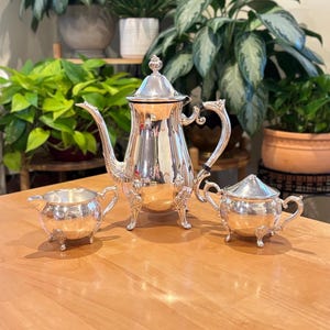 May include: A silver tea set with a teapot, creamer, and sugar bowl. The teapot has a curved spout and ornate handle. The creamer and sugar bowl have matching handles and lids. All pieces have decorative feet.