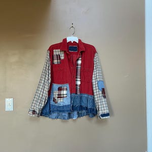 May include: A patchwork jacket with a red body, plaid sleeves, and denim and plaid accents. The jacket has a ruffled denim hemline and is hanging on a white hanger.