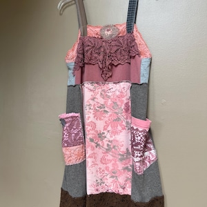 May include: A sleeveless dress featuring a patchwork design with various fabrics in shades of pink, gray, and brown. The garment includes a lace-trimmed bodice, two side pockets with lace overlays, and a dark brown lace hemline at the bottom.