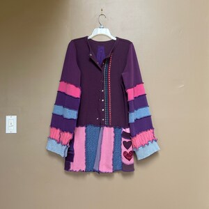 May include: A purple, pink, and blue patchwork cardigan with a button-up front and long sleeves. The cardigan has a heart-shaped patch on the front.