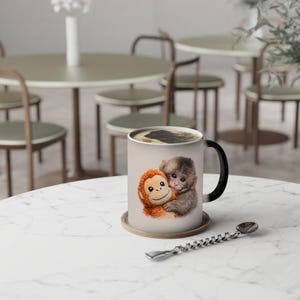 Könnte beinhalten: Eine Keramik-Kaffeetasse mit schwarzem Henkel und beigefarbenem Äußeren. Die Tasse zeigt eine Illustration von zwei sich umarmenden Affen. Die Tasse steht auf einem runden Untersetzer auf einem weißen Marmortisch.