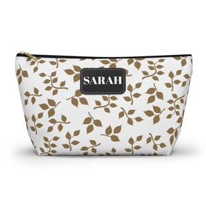 May include: A white cosmetic bag with a repeating pattern of brown leaves and branches. The bag has a black leather patch with the name "SARAH" in white letters. The bag has a gold zipper.