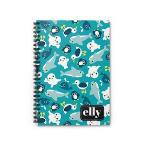 May include: Spiral-bound notebook with a teal cover featuring a pattern of cartoon sea creatures, including penguins, seals, and sharks. The notebook has a black spiral binding and the name "elly" is printed on a black and white label.