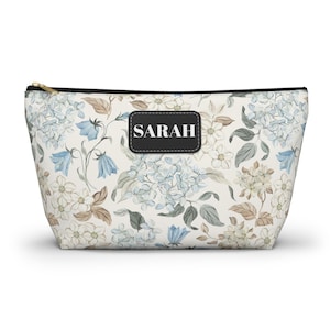 Puede incluir: Un neceser con estampado floral y cremallera dorada. La bolsa presenta un estampado de hortensias azules y blancas, con el nombre "SARAH" en una etiqueta negra. La bolsa está diseñada para guardar maquillaje y otros productos de belleza.