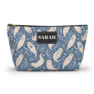 Puede incluir: Un estuche con cremallera que presenta un patrón repetitivo de nutrias de color beige claro nadando entre algas y conchas azules. Un parche rectangular negro está centrado en el frente con el nombre SARAH impreso en letras blancas.