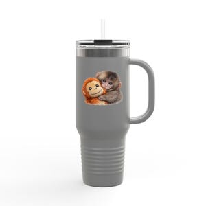 May include: Grey travel mug with a handle and a straw. The mug features an illustration of two monkeys hugging. One monkey is brown and the other is grey. The mug has a silver rim and a black lid.