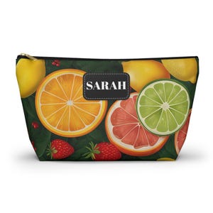 Può includere: Un beauty case con cerniera con un vivace motivo a frutta. Il design include fette di arancia, limoni, pompelmi e fragole su uno sfondo verde scuro. Il nome "SARAH" è visualizzato su un'etichetta in bianco e nero.