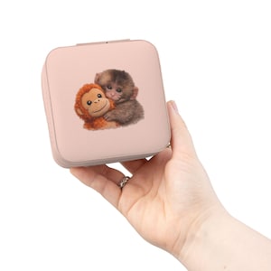 May include: A light pink square jewelry box featuring a graphic of two hugging monkeys. The box is held in a hand against a white background. The monkeys are brown and orange with detailed fur.