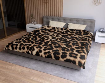 Puede incluir: Una cama cubierta con una manta con estampado de leopardo. La manta presenta un patrón de manchas negras sobre un fondo marrón claro. La cama tiene un cabecero gris y dos almohadas blancas. La habitación tiene suelo de madera clara y una alfombra gris.