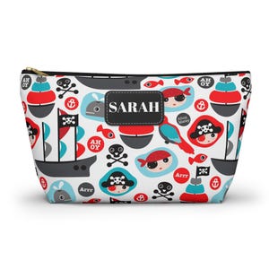 Pode incluir: Uma bolsa branca com zíper com um padrão de tema pirata. O design inclui navios piratas, caveiras, peixes vermelhos e rostos de piratas. O nome "SARAH" é exibido em um patch preto. A bolsa tem um zíper dourado.