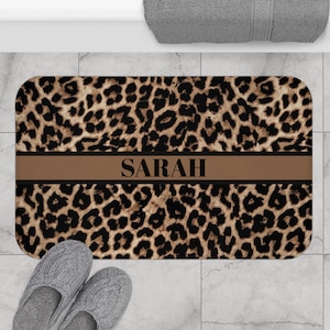 May include: A rectangular bath mat with a leopard print design. A brown band with the name "SARAH" in black lettering is centered on the mat. Two gray slippers are at the bottom of the mat.