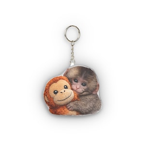 Pode incluir: Um porta-chaves com uma imagem comovente de dois macacos a abraçar-se. Um é um brinquedo de pelúcia em tons de laranja, e o outro é um macaco real com pelo castanho. O porta-chaves tem um anel e uma corrente prateados.