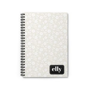 Könnte beinhalten: Ein Spiralnotizbuch mit einem weißen Blumenmuster auf hellbeigem Hintergrund. Das Notizbuch hat das Wort "elly" in einem schwarzen Rechteck unten rechts. Die Spiralbindung ist schwarz.