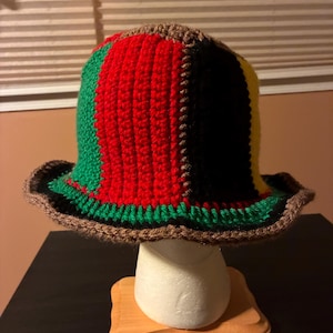 May include: A handmade crochet bucket hat featuring vertical panels in vibrant red, green, black, and yellow yarn. The hat is finished with a brown crocheted trim along the edge of the brim and a brown crocheted detail at the crown.