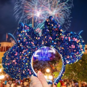 May include: A blue sequined headband with large mouse ears. The ears and bow are covered in iridescent sequins. The headband is held up against a backdrop of fireworks and a crowd of people.