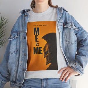 Peut inclure: Un t-shirt gris avec un motif graphique représentant une silhouette de casque spartiate sur fond orange. Le texte "ONLY ONE WINS" est au-dessus des mots "ME VS ME". La personne porte une veste en jean bleue.