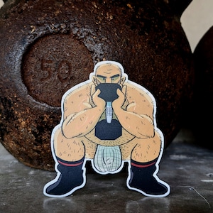 Può includere: Un adesivo con un'illustrazione a cartoni animati di una figura muscolosa e calva che solleva un manubrio. La figura indossa stivali neri con strisce rosse e un perizoma. Lo sfondo include un kettlebell marrone scuro arrugginito.