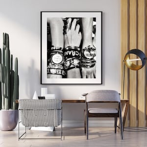 Puede incluir: Fotografía en blanco y negro de tres muñecas adornadas con múltiples pulseras y relojes. Los relojes tienen esferas redondas y correas metálicas. Algunas pulseras tienen texto. La imagen está enmarcada y colgada en una pared blanca.