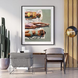 Op de afbeelding: Een ingelijste kunstdruk toont drie figuren die zonnebaden op witte handdoeken. De figuren dragen badkleding in geel, roze en turquoise. Het kunstwerk wordt tentoongesteld aan een witte muur boven een houten bureau.