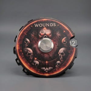 May include: A round, black and red game piece with the word "WOUNDS" at the top. The center features a silver disc and a skull design. The number "12" is visible on the right side. This game accessory is designed for tabletop gaming.