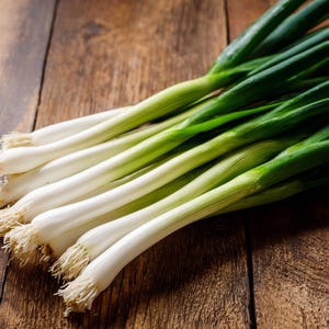 May include: A close-up shot of fresh green onions, also known as scallions, arranged on a wooden surface. The onions have white bulbs and vibrant green stalks. The wooden surface has a natural, warm tone.