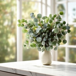 May include: A bouquet of faux eucalyptus in a cream-colored vase. The eucalyptus has round, green leaves and reddish-brown stems. The vase sits on a white marble countertop, with a blurred window in the background.